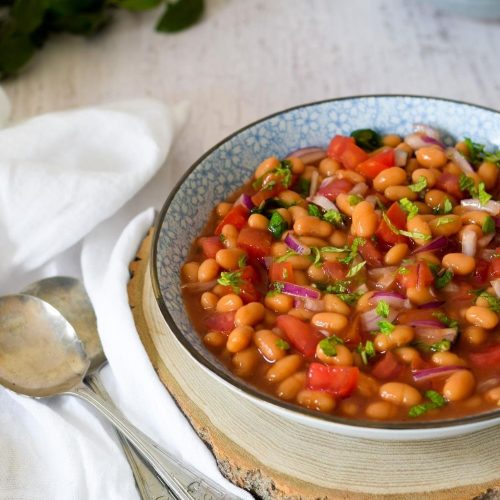 Baked Bean salad in a blue bowl on a piece of wood with spoons on left