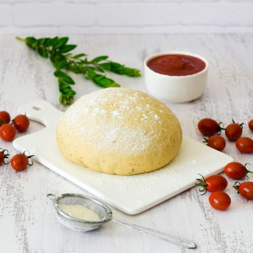 Piza dough on a white board with pizza sauce at the back and tomatoes placed around