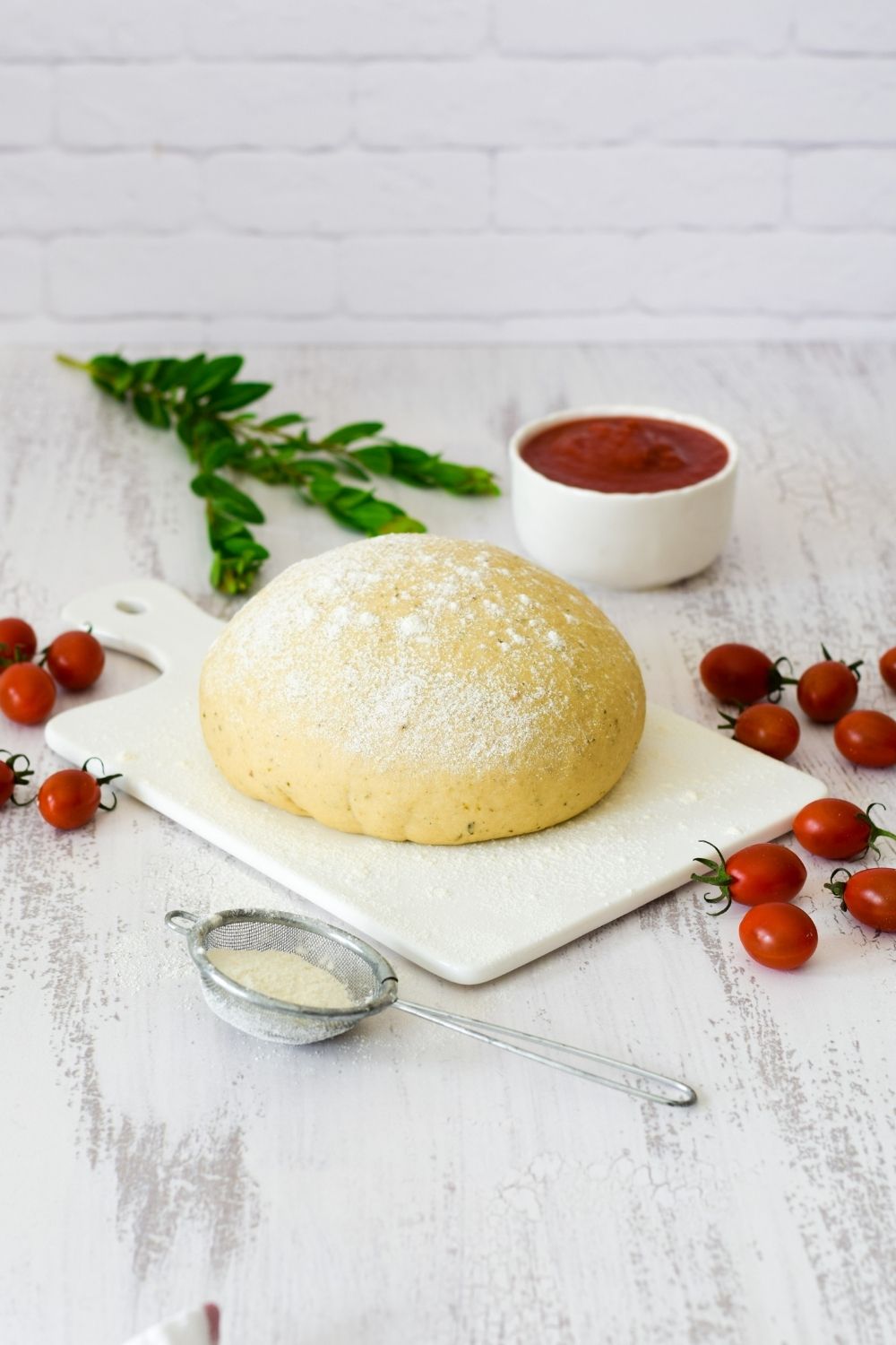 Pizza dough on a white board with pizza sauce at the back and tomatoes placed around