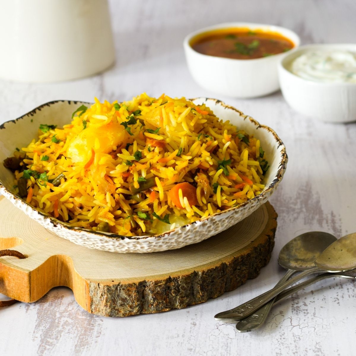 Vegetable biryani in a white bowl on a piece of wood with dal and raita at the back with spoons on right