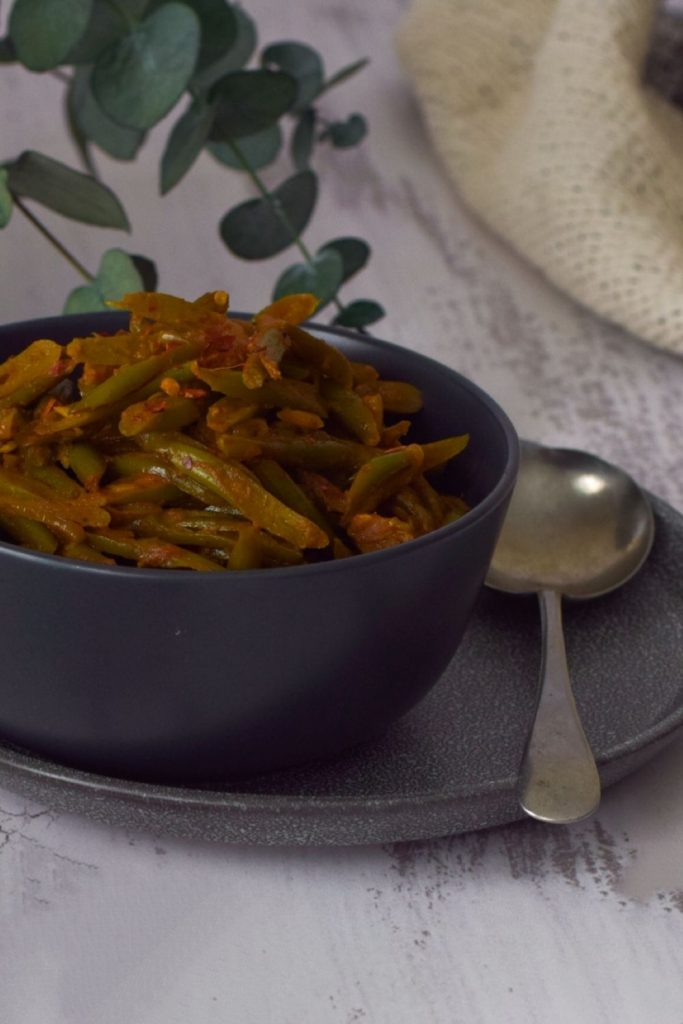 Green Bean Curry in a black bowl with spoon on right, leaves at left back