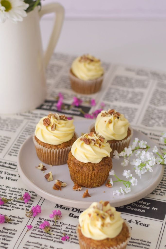 Carrot Cupcales on a white plate with pink flowers around and a vase back left