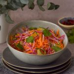 Carrot salad in a bowl with chilli at the back right and green leaves on the left