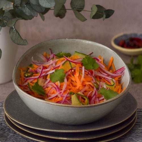 Carrot salad in a bowl with chilli at the back right and green leaves on the left