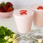 Strawberry Lassi in 2 glasses with flowers on the left and strawberries at the back