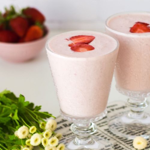 Strawberry Lassi in 2 glasses with flowers on the left and strawberries at the back