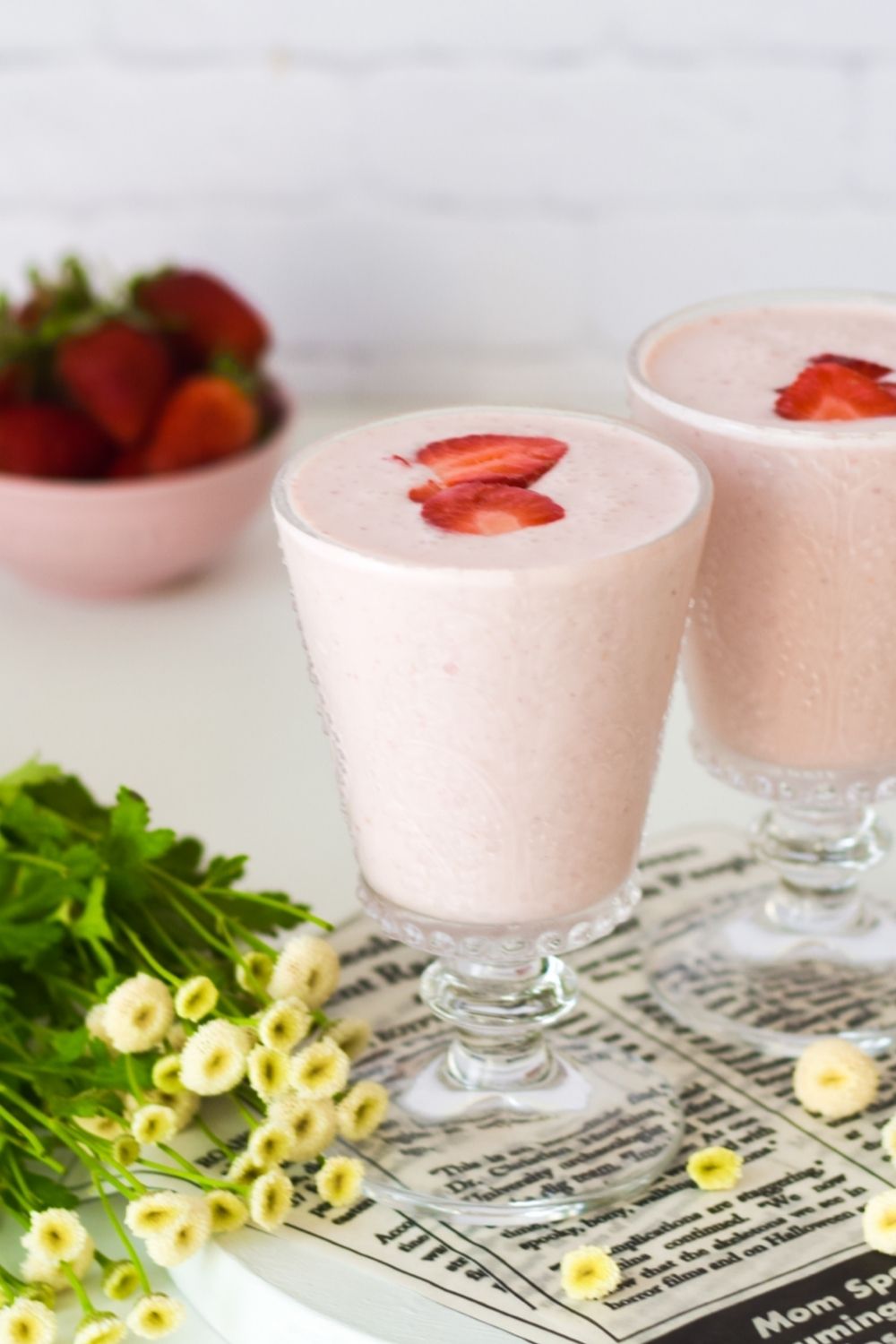 Strawberry Lassi in 2 glasses with flowers on the left and strawberries at the back