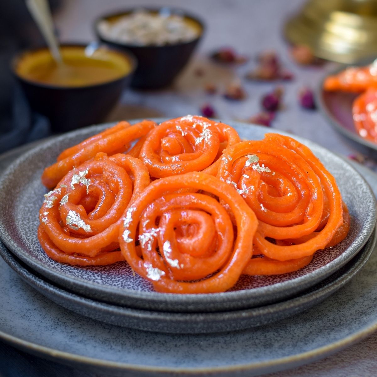 Jalebi in on stacked grey plates with cardamom and syrup back left and more jalebi on right