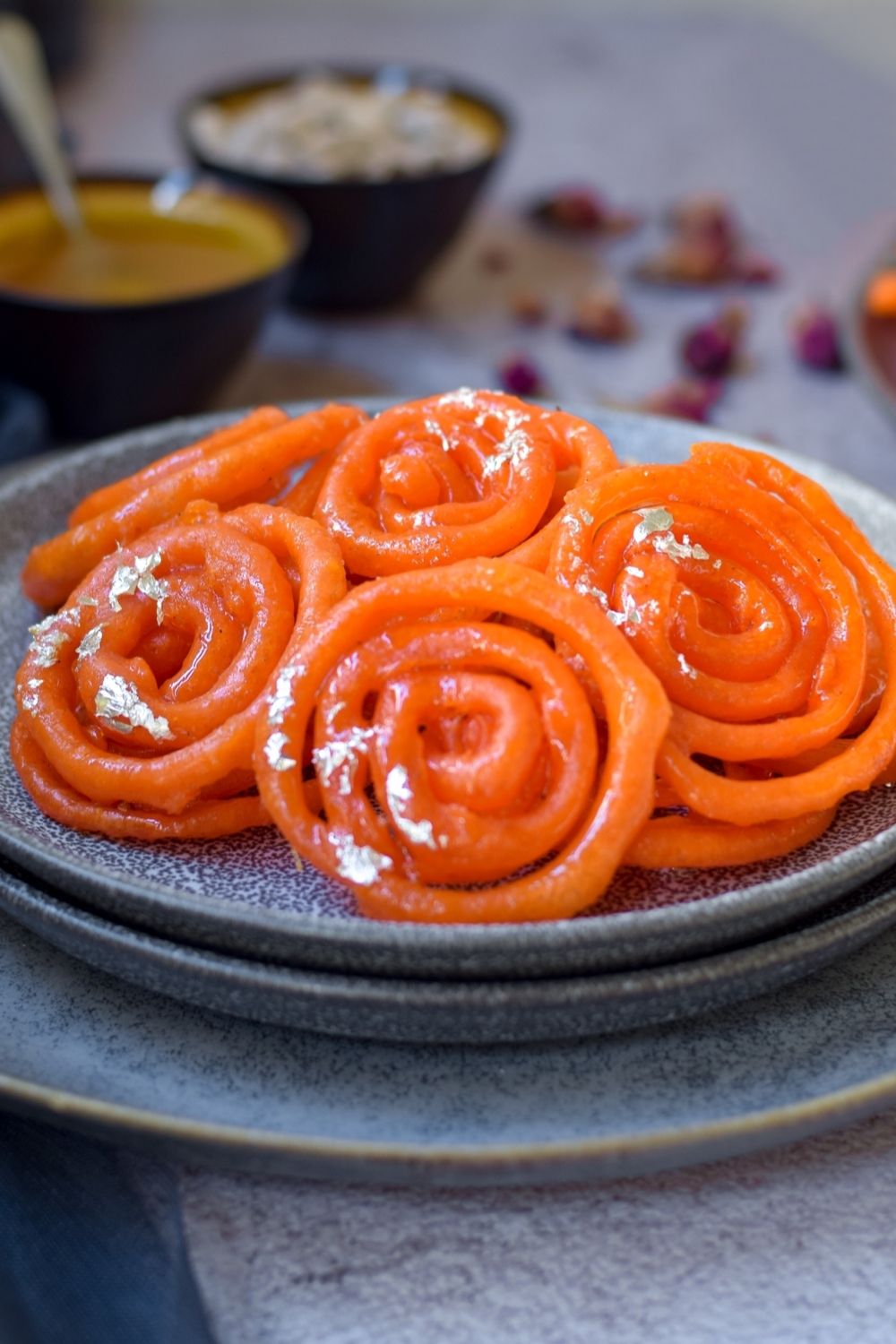 Jalebi in on stacked grey plates with cardamom and syrup back left and more jalebi on right