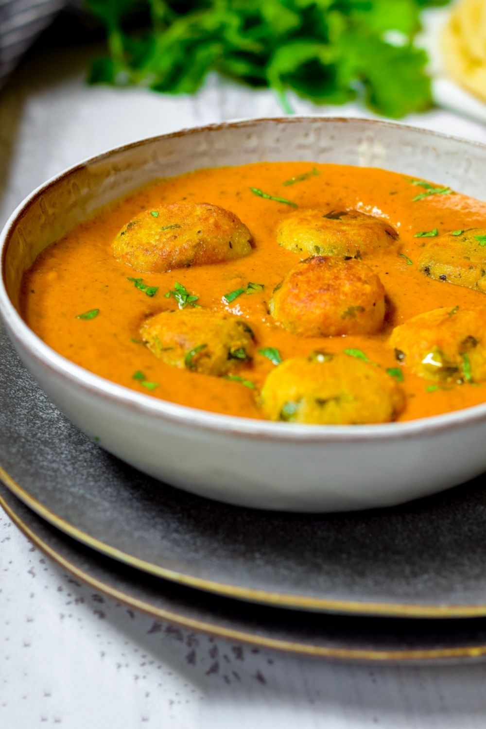 Malai Kofta in a white bowl on top of black plates with coriander at the back