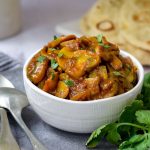 Mushroom Curry in a white bowl with naan bread back right and spoons on left
