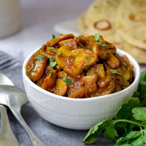 Mushroom Curry in a white bowl with naan bread back right and spoons on left