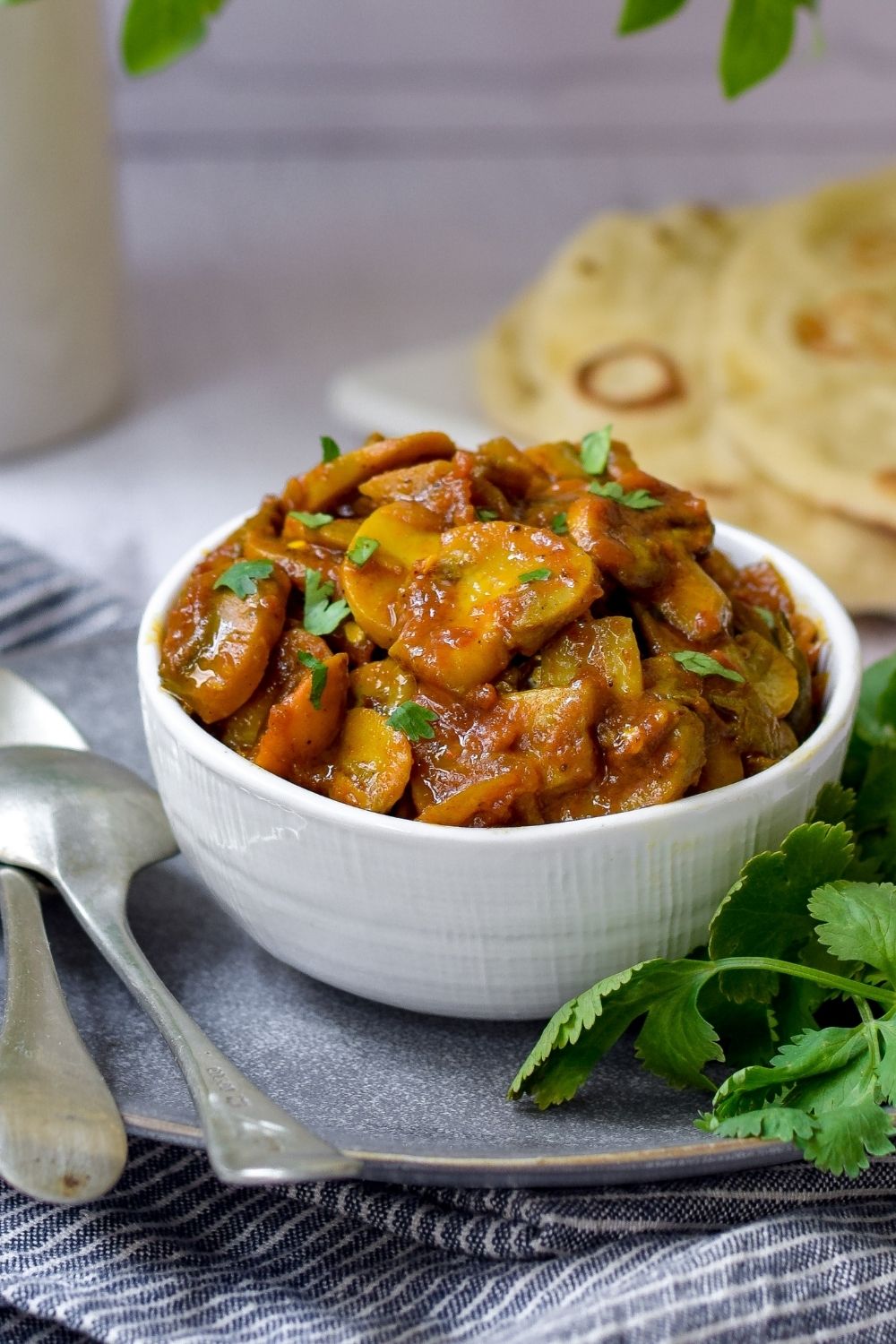 Mushroom Curry in a white bowl with naan bread back right and spoons on left