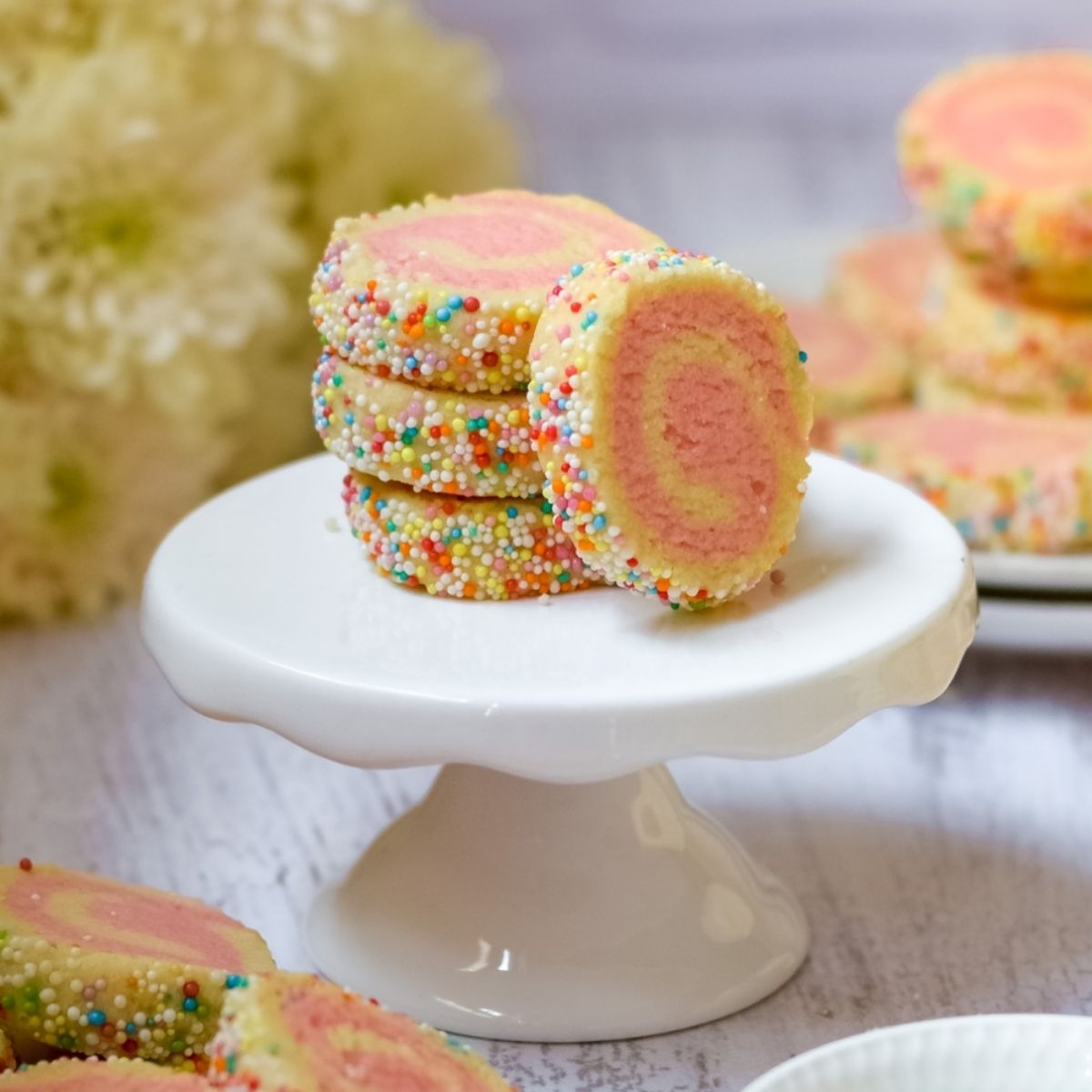Pinwheel cookies on a cake stand with more on the right and flowers on the left