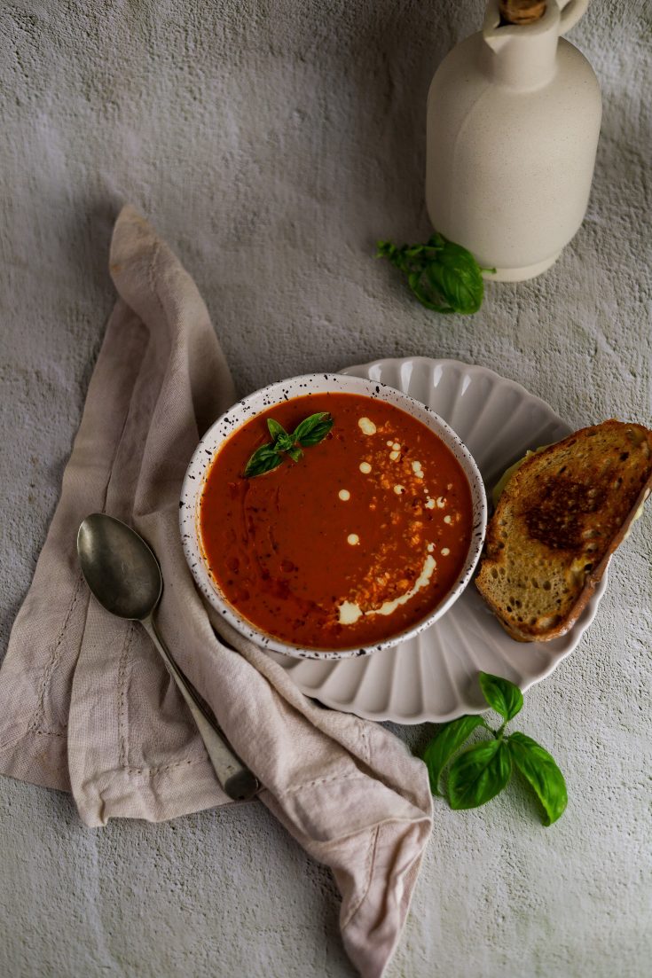 Roasted red pepper and tomato soup in a white bowl with a spoon on the left and grilled cheese sandwich on the right