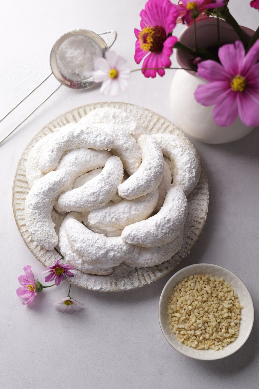 Kourabiedes in a cream coloured plate with a sieve on top left and almonds bottom right